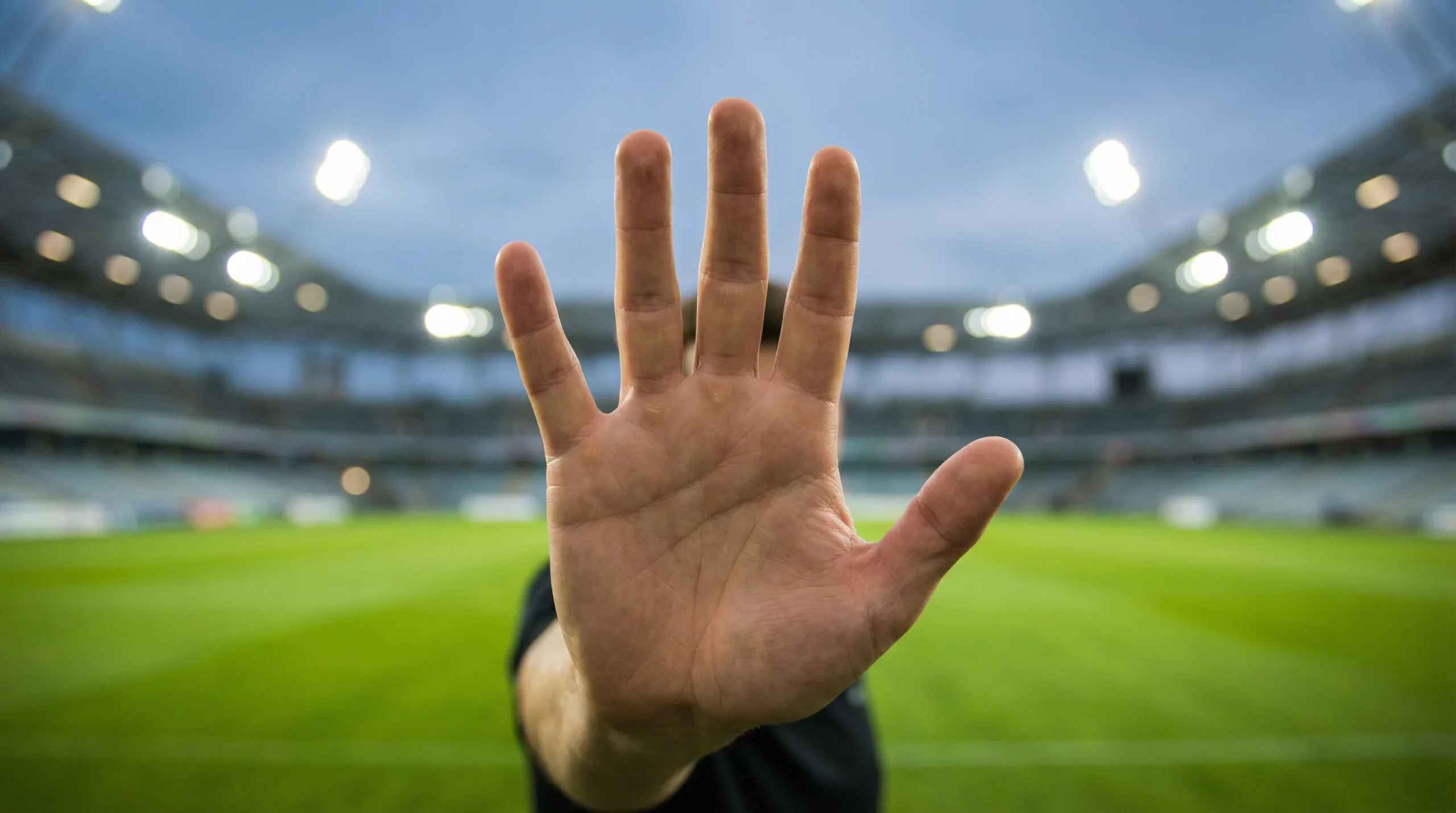 Mano levantada en señal de pausa frente a un campo de fútbol iluminado al fondo